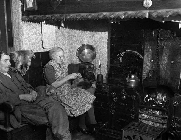 Delwedd ddu a gwyn yn dangos dau berson yn eistedd wrth y tân gyda chath. Mae'r deunyddiau ar gyfer y canhwyllau yn sychu uwchben y stôf wrth ymyl tân agored.